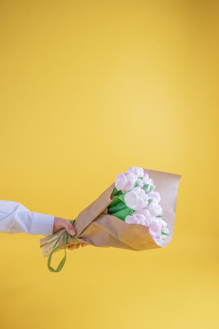 A person wearing a white shirt holds a bouquet of fresh, pale pink tulips with green stems, wrapped in light brown kraft paper and secured with a green ribbon. The bouquet is positioned horizontally a