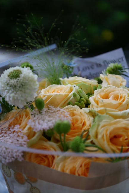 A close-up of a floral arrangement featuring pale peach roses with soft, layered petals, complemented by delicate white and green blooms, including fluffy white pom-pom chrysanthemums and small green 