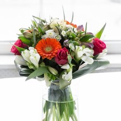 This radiant bouquet, a true testament to professional floristry in Muswell Hill, bursts with exuberant colour and detailed craftsmanship. The eye is immediately drawn to large, jubilant gerbera daisies-two in bold orange and two in soft peach-each anchored by a velvety, chocolate brown centre. They're offset by luxurious, deep fuchsia roses, their tightly furled petals suggesting romance and refined beauty, much like those found in bloom near Broadway's leafy avenues. Pure white alstroemeria and small spray roses serve to brighten and soften the overall aesthetic, imparting a luminous quality similar to early morning light breaking across Alexandra Palace gardens. Subtle touches of blush pink blooms and airy white accents contribute to a layered effect, filling the bouquet with depth and vitality. The arrangement's base is skillfully enveloped in sculpted, broad aspidistra leaves, whose glossy green expanse curves gently to provide both support and a stunning visual anchor. Slender green shoots and textural foliage fill the ensemble, enhancing its fullness and giving a nod to Muswell Hill's lush environment. Bathed in diffuse, natural light-perhaps streaming through a North London window-the bouquet radiates positive energy and heartfelt sentiment. This creation is perfect for commemorating life's moments, from joyful celebrations to thoughtful gestures of appreciation, and showcases the high standards and caring touch of local Muswell Hill florists.